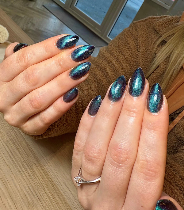 Close-up of hands with cat eye magnetic gel in blue and green metallic nail polish on a wooden surface.